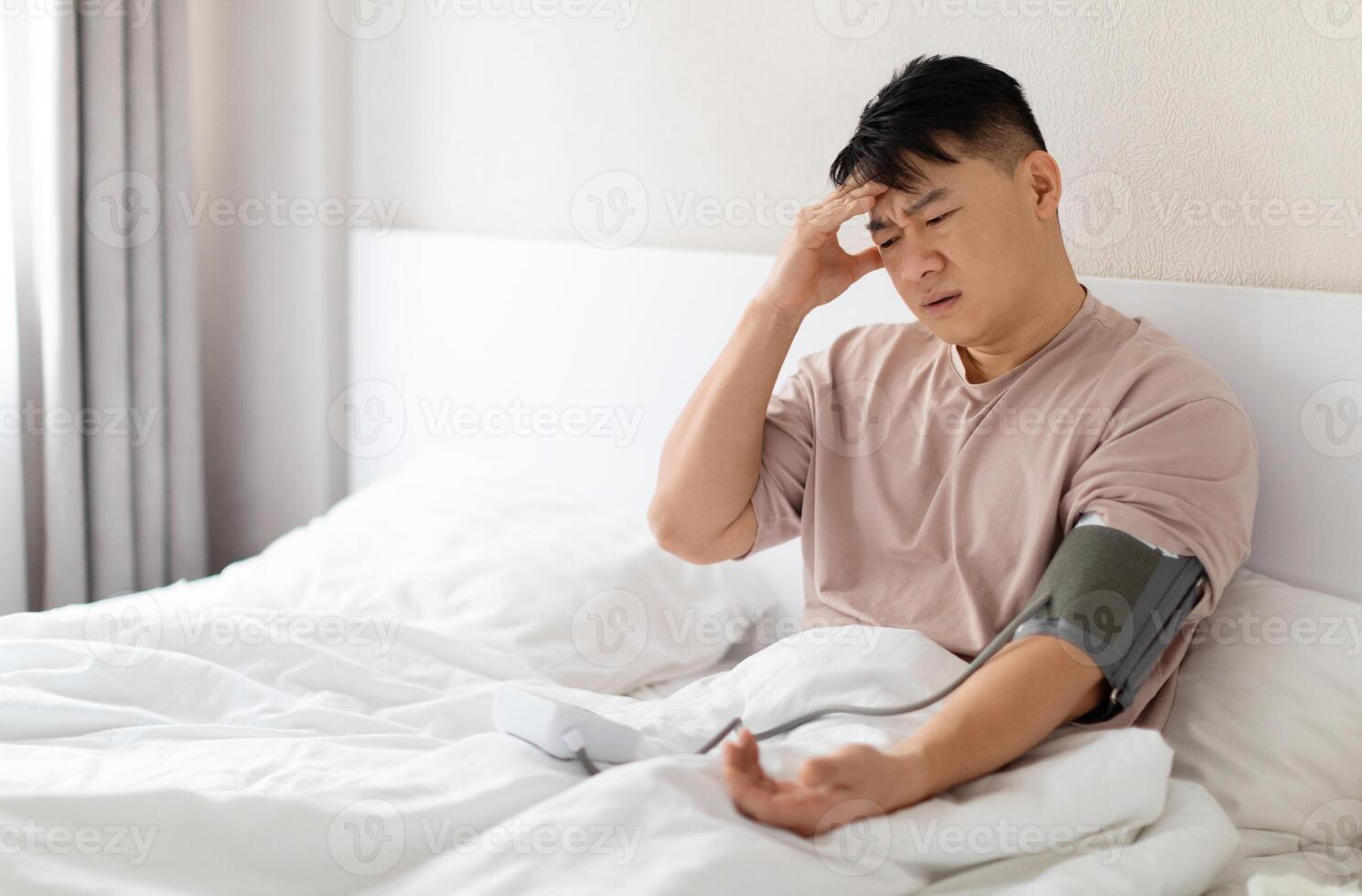 Unhealthy asian man in pajamas sitting in bed at home, checking blood pressure on his own. Sick male using modern tonometer, experiencing hypertension caused by stress, touching head, copy space photo