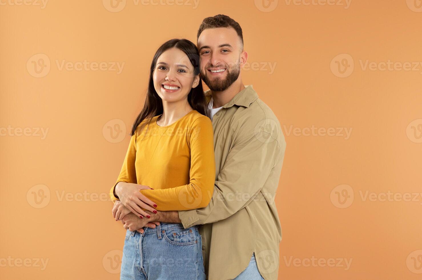 Happy young caucasian man hugging his girlfriend posing on orange background in studio, smiling to camera. Millennial family couple embracing for photo. Love and romantic date, Valentine's day photo