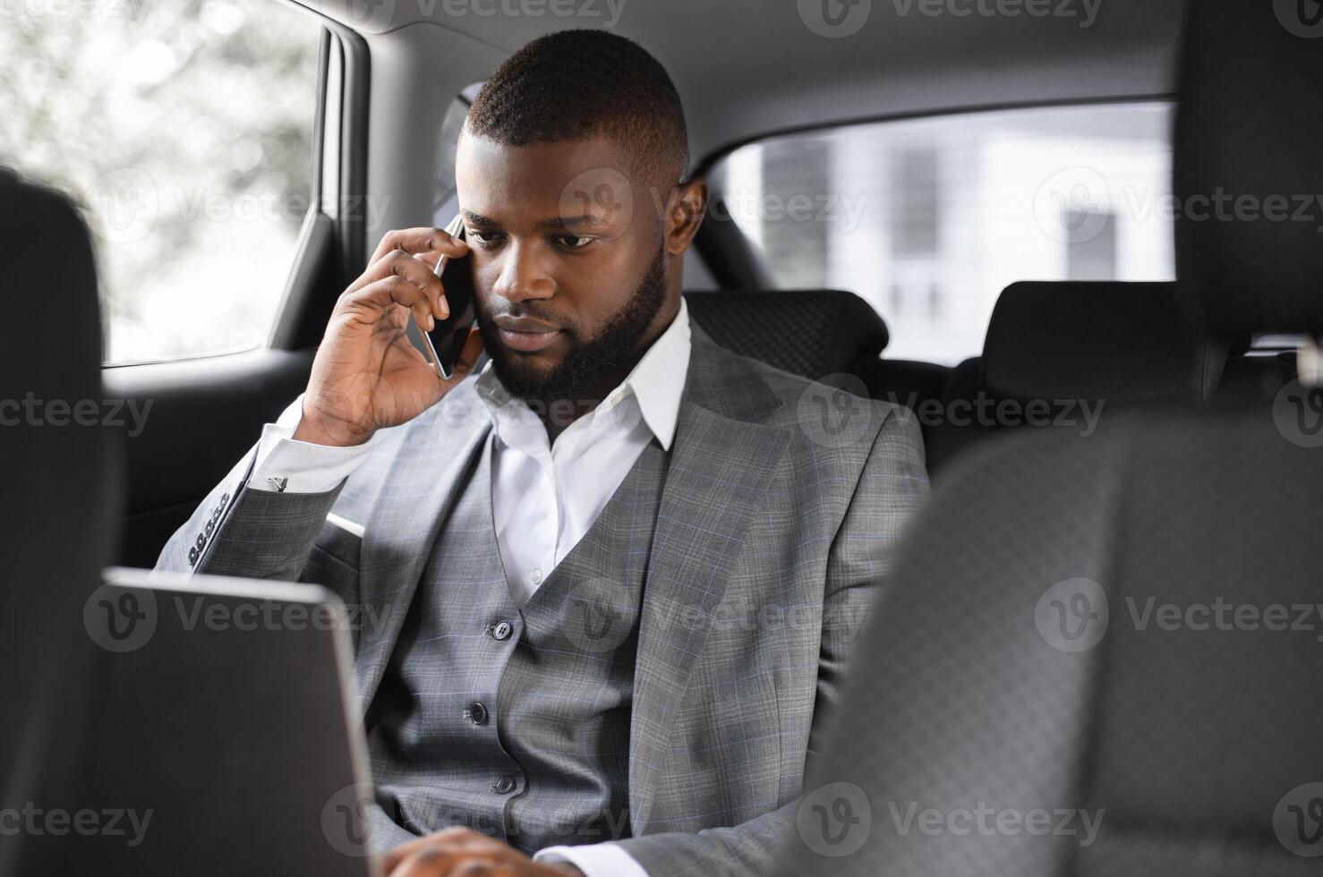 Concentrated black businessman talking on mobile phone and looking at laptop screen while going to business meeting by car, young african manager sitting on back seat in car and working, closeup photo