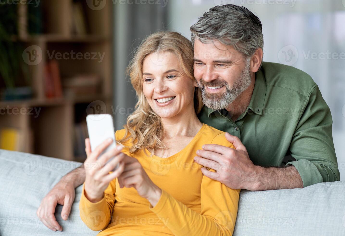 Portrait Of Smiling Middle Aged Couple Resting With Smartphone On Couch, Happy Mature Spouses Watching Photos Or Browsing New Mobile App While Relaxing In Living Room, Closeup Shot With Free Space