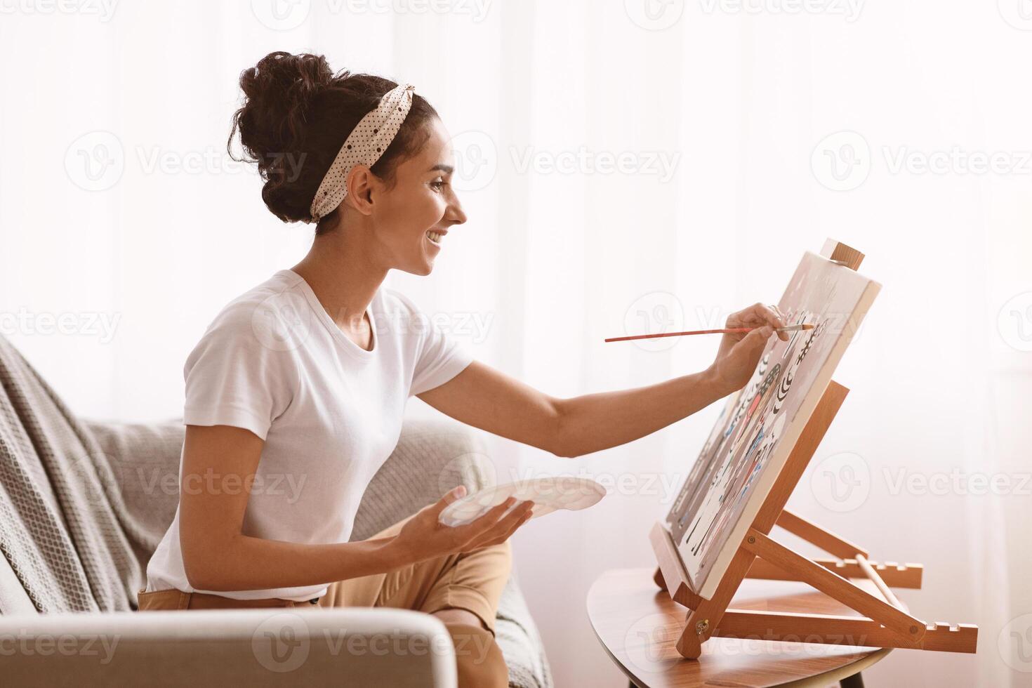 Side view of happy young woman sitting on couch and painting, using palette, brush and easel, home interior. Creative lady enjoying new hobby, painting still life, copy space. Entertainment concept photo