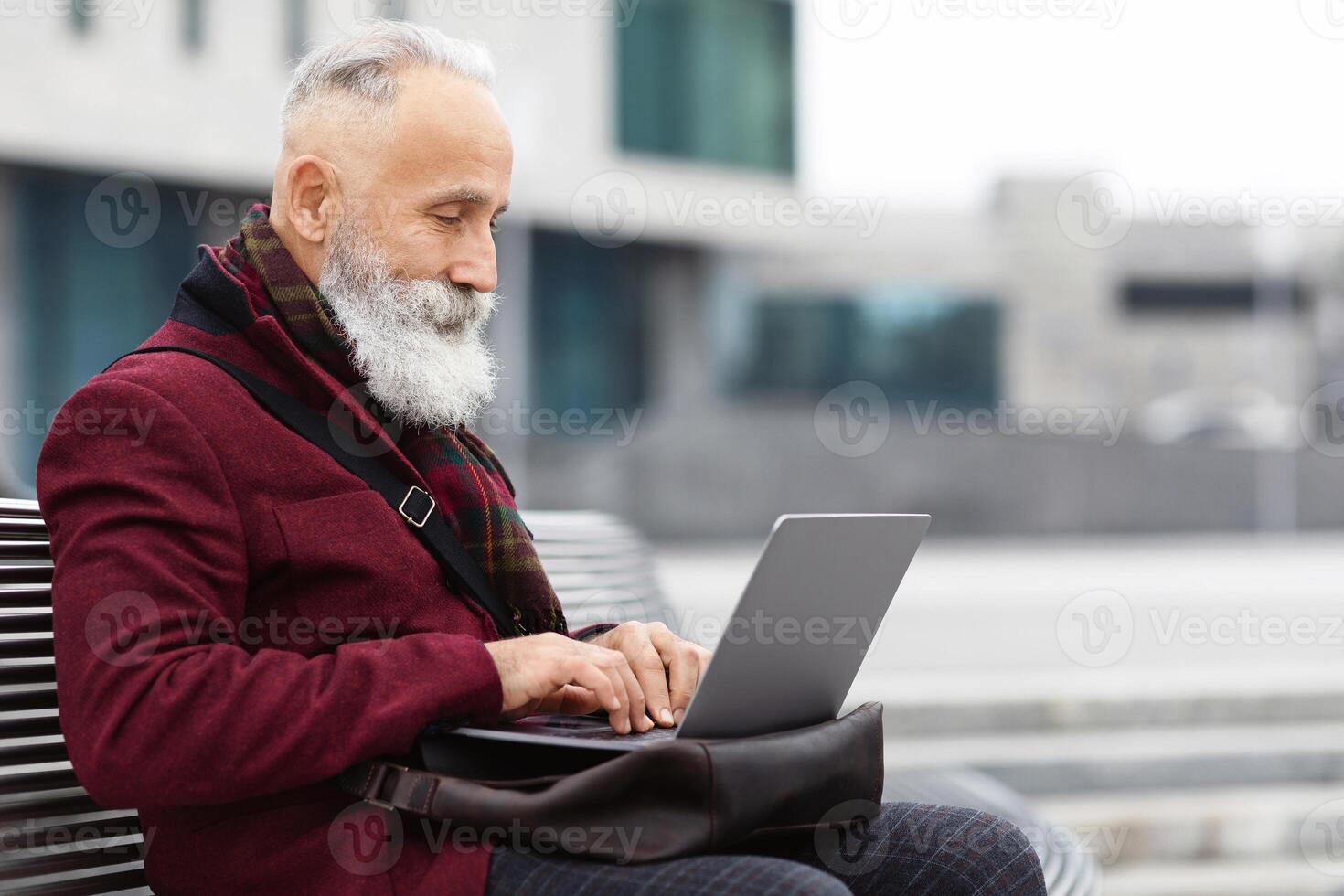 Senior bearded businessman with modern laptop sitting on bench, typing on keyboard, sending email, empty space. Stylish aged grey-haired man sitting on the street by business center, working online photo