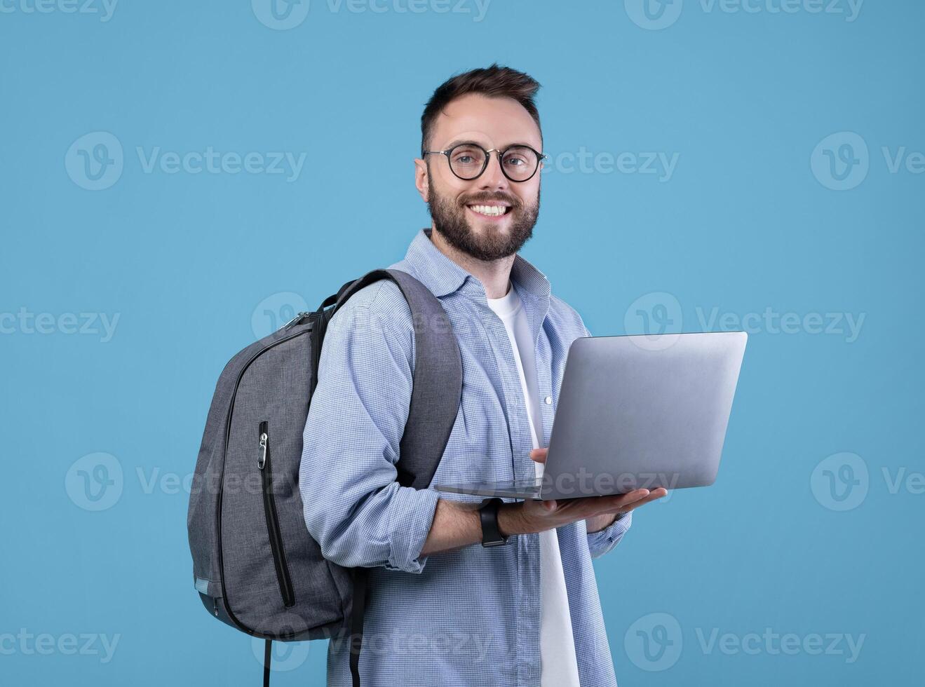 Smart millennial student with backpack using laptop computer over blue studio background. Cheerful young guy doing online work, communicating with family or friend, attending webinar photo