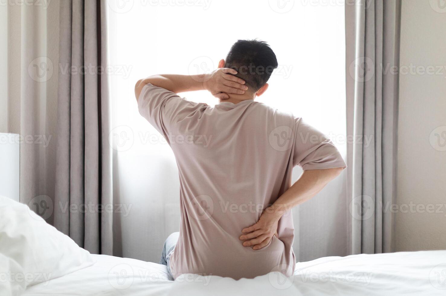 Tired man suffering from back pain after sleeping in bedroom, waking up in morning, sitting on bed and touching lower back and neck, experiencing body aches, wearing pajamas, copy space, back view photo