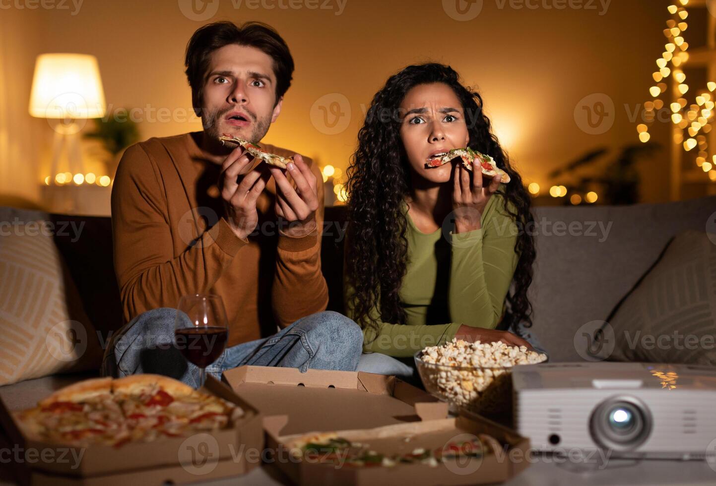 Shocked Couple Watching Horror Movie And Eating Pizza Using Home Cinema Projector Sitting On Sofa In Living Room Indoors At Night. Family Entertainment And Weekend Leisure Concept photo