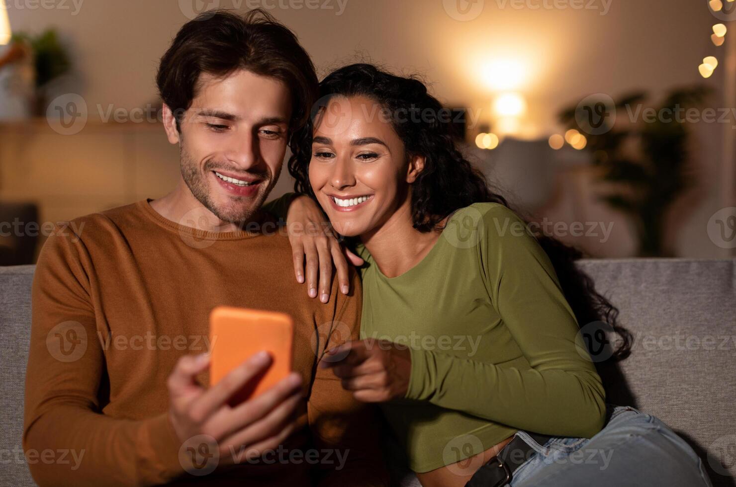 Cheerful Couple Using Cellphone With New Application, Wife Pointing Finger At Phone Browsing Internet Sitting On Couch At Home. Spouses Recommending Great App. Gadgets Concept photo