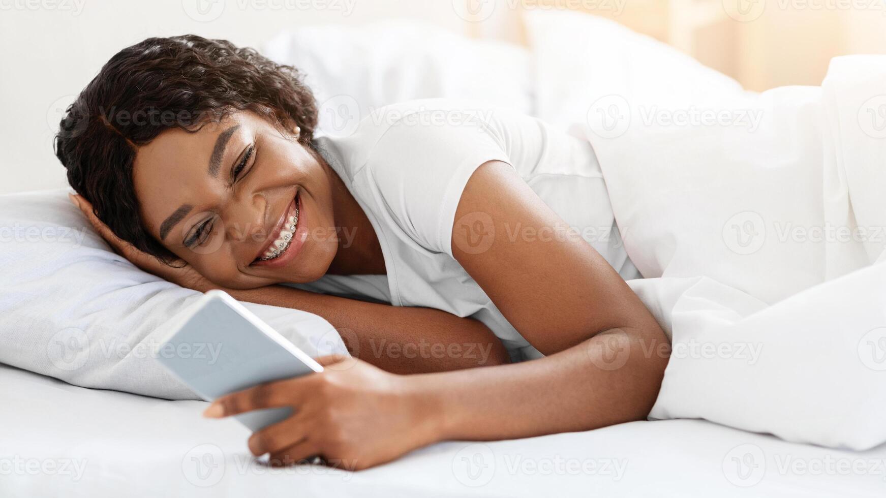 Happy young black woman using smartphone in bed before sleeping, chatting with her lover or girlfriends. Cheerful african american lady checking social media while laying in bed, copy space photo