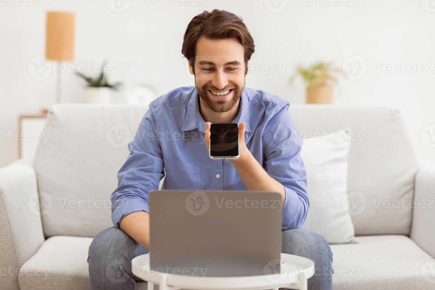 Cheerful Guy Using Voice Assistant On Mobile Phone Or Talking Using Loudspeaker Mode On Smartphone Sitting At Laptop Computer On Sofa At Home. Voice Search Application, Gadgets Lifestyle. photo