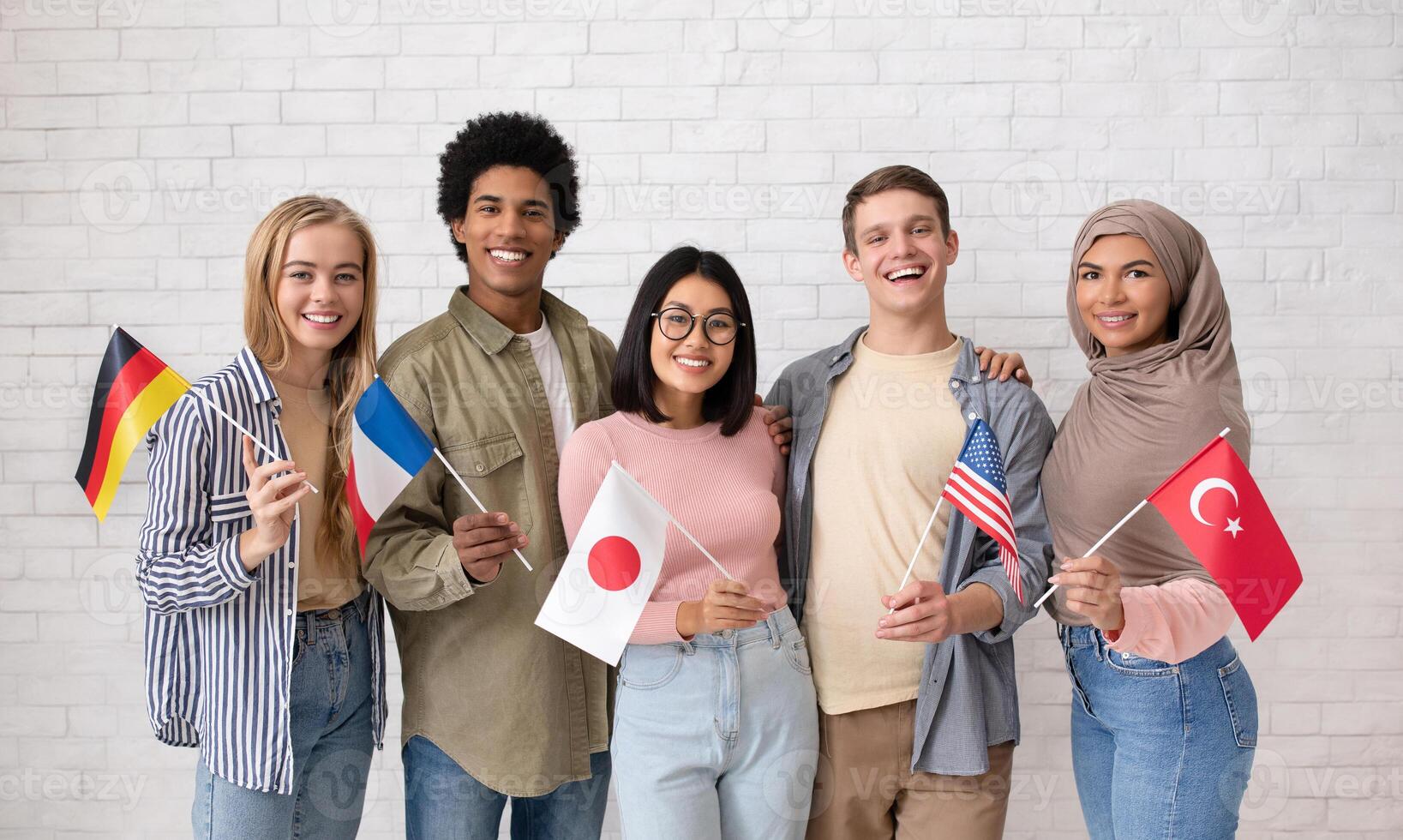 Student exchange, international friendship. Multiethnic young students with flags of different countries ready to study and look at camera on brick white wall background, studio shot, free space photo