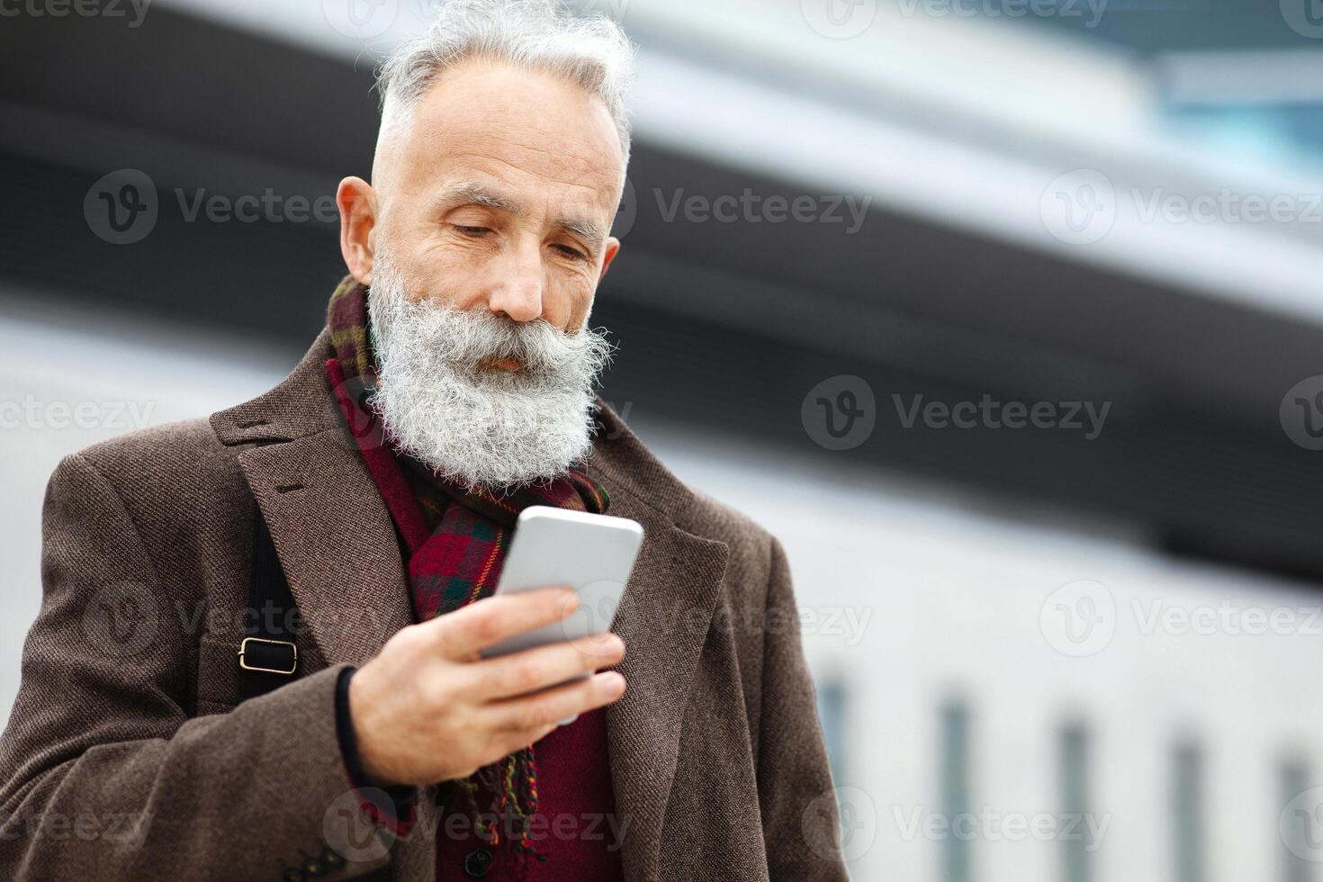 Grey-haired elderly businessman using smartphone on the street, checking his agenda, copy space. Bearded aged man using mobile phone, websurfing or chatting with colleagues, having lunch break photo
