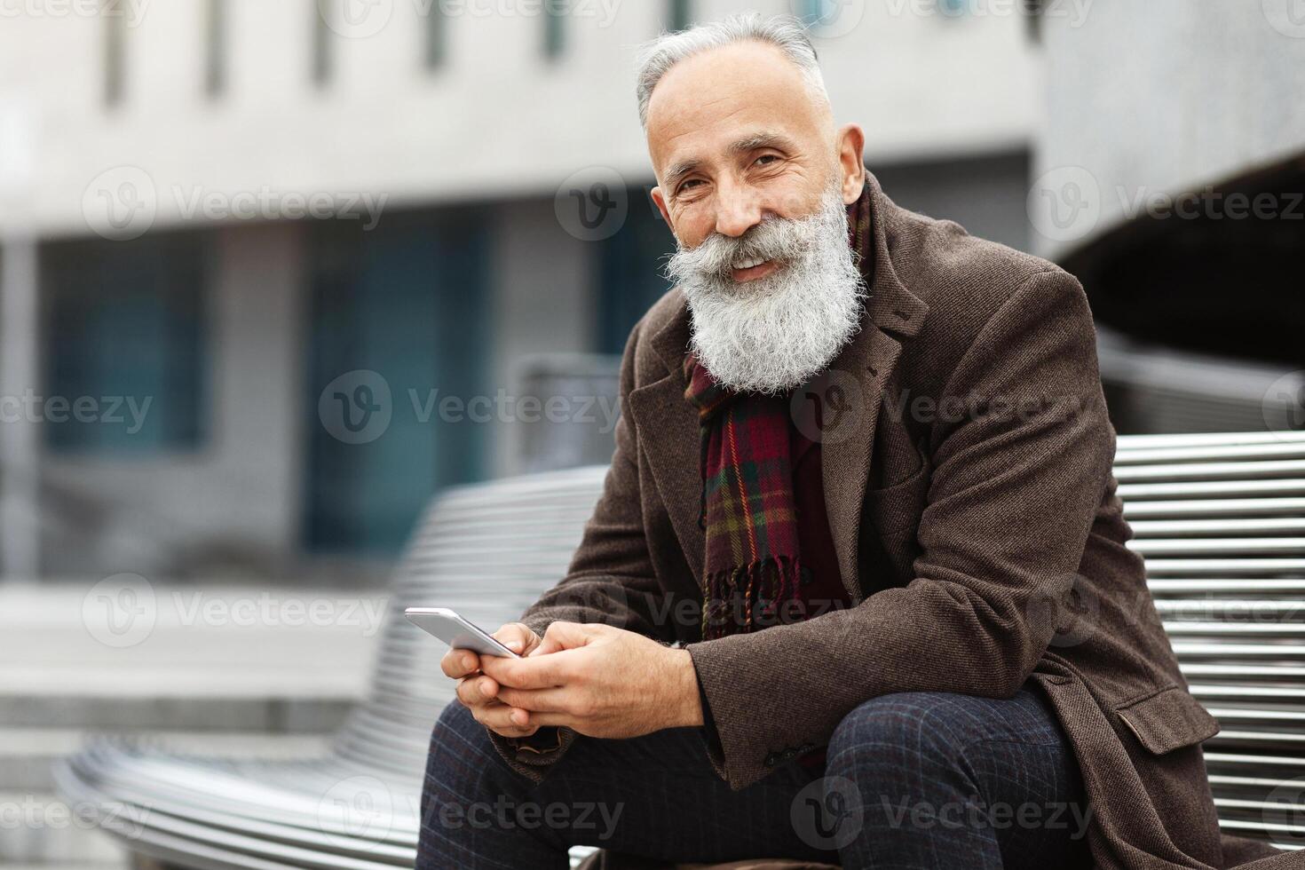 Smiling senior grey-haired man with mobile phone sitting on bench on street, smiling at camera, copy space. Good-looking aged man with long beard resting by office building, having lunch break at job photo