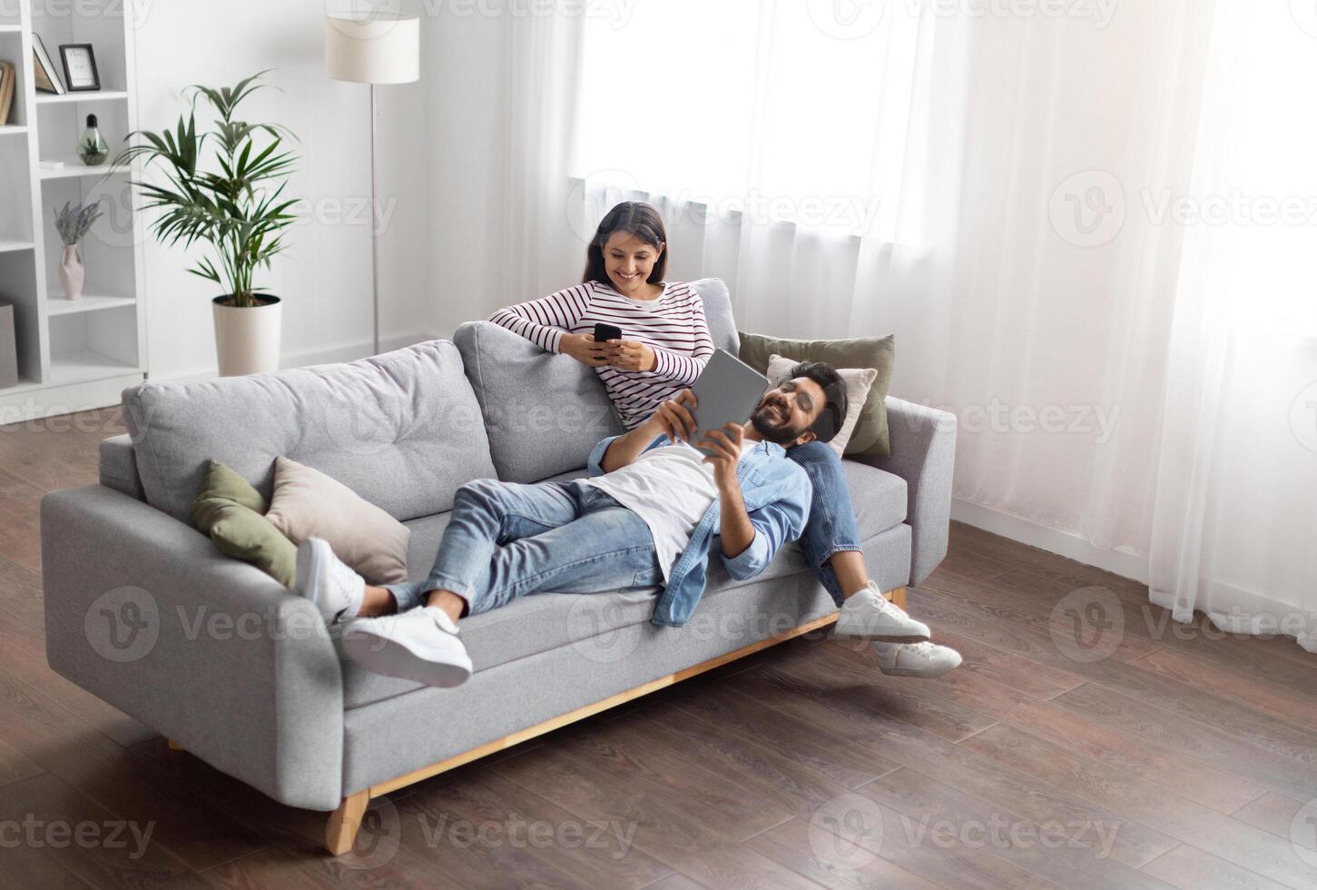 High angle view of relaxed cheerful stylish millennial eastern lovers man and woman chilling on couch in cozy living room, using gadgets at home, scrolling on social media, reading online, copy space photo