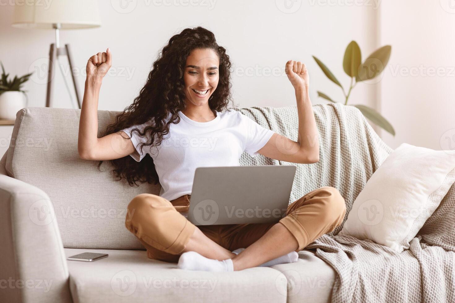 Emotional young woman with dark curly hair looking at laptop screen and raising hands up, freelancer working from home, sitting on couch and using modern laptop, blogging or copywriting, copy space photo