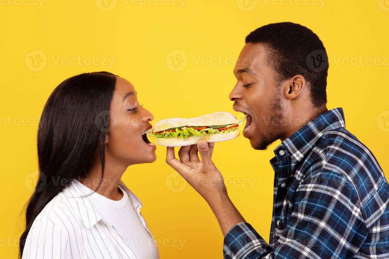 Romantic date and fast food. Cute millennial african american man and woman eat one sandwich for two at covid-19 quarantine, isolation on yellow background, studio shot, profile, copy space photo