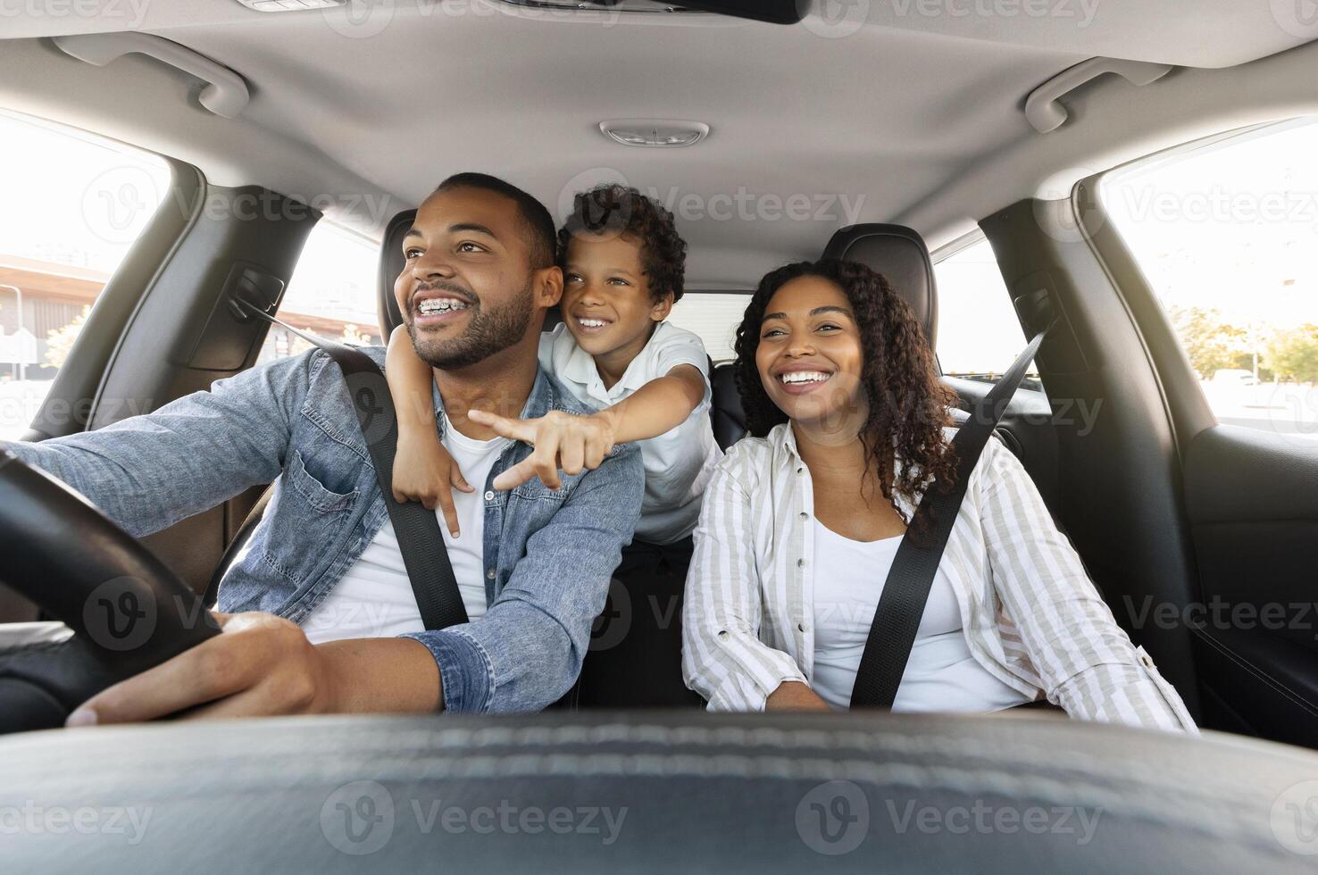 Beautiful cheerful loving african american family enjoying car trip. Cute preteen black boy embracing his father driving auto and pointing at road, happy parents and child spend weekend together photo