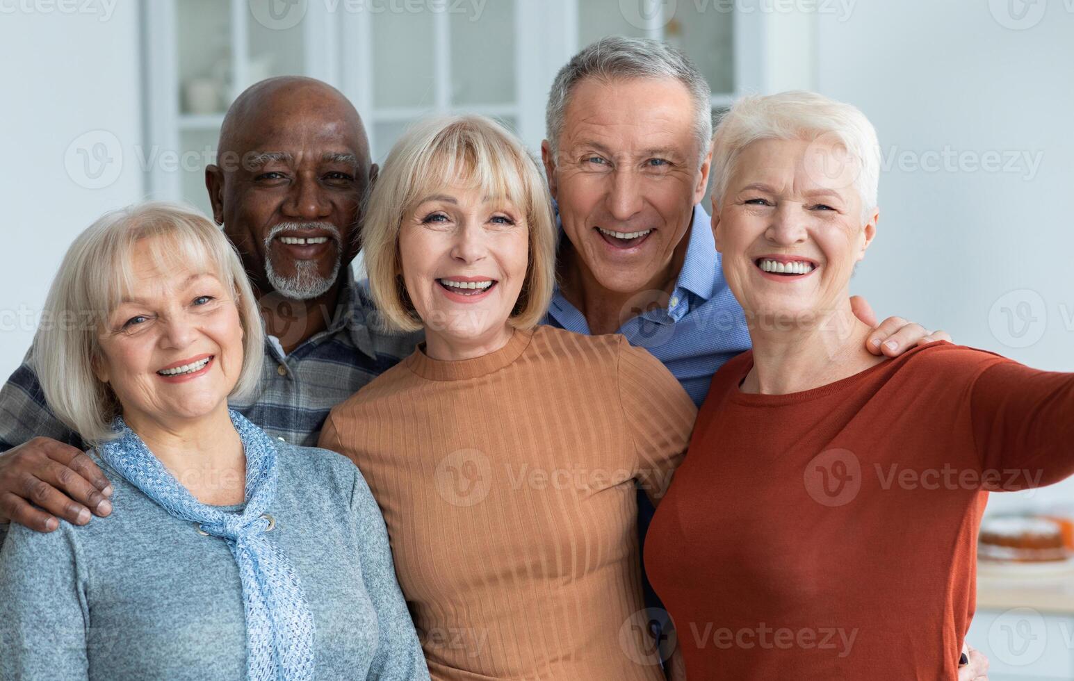 Multiracial group of cheerful positive senior people men and women in casual outfits taking selfie together while having home party, hugging and smiling at camera, kitchen interior photo