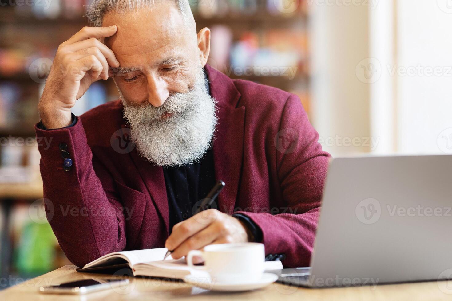 Thoughtful aged businessman with grey hair checking agenda, taking notes in notebook, working with laptop at cafe, copy space. Elderly concentrated man looking for job, checking his list, drinking tea photo