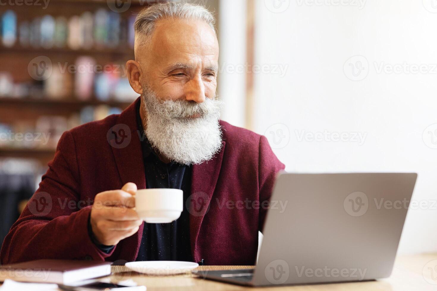 Positive senior bearded man with grey hair drinking coffee and using laptop at cafe, copy space. Stylish aged businessman in burgundy jacket enjoying his tea while working online photo