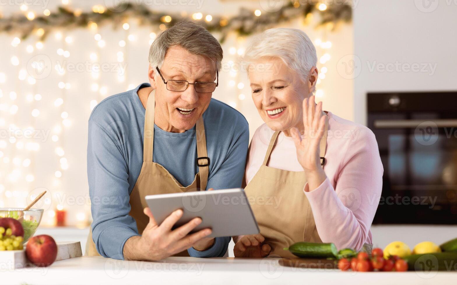 Joyful Senior Couple Using Tablet Making Call Standing In Kitchen On Christmas Eve. Remote Xmas Celebration Online. Older Spouses Chatting With Distant Family Using Computer photo