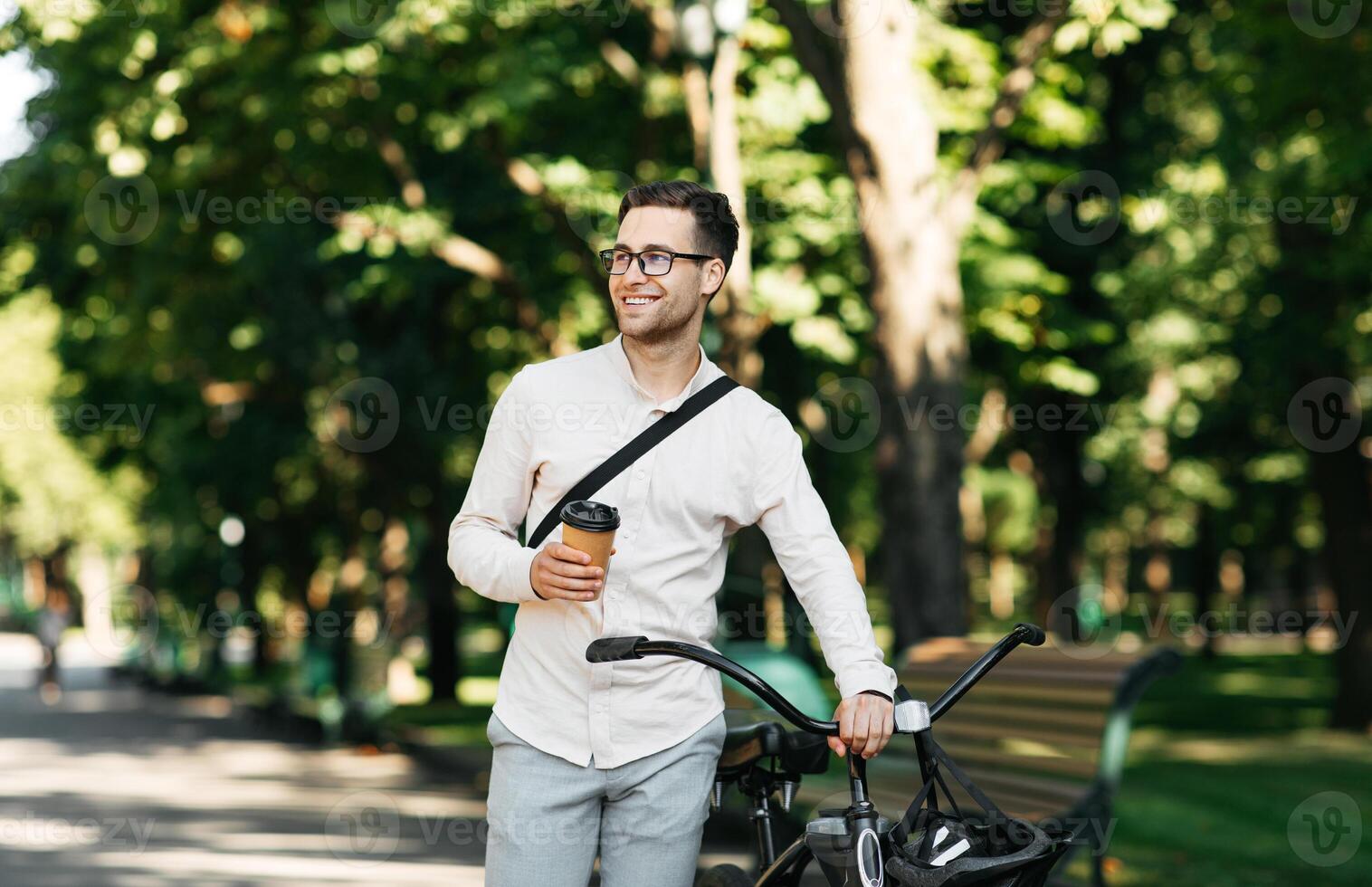 Healthy and active lifestyle, going to work on eco transport. Smiling attractive millennial male employee in glasses with bag and cup of takeaway coffee, moving bike in city park in summer, outdoor photo
