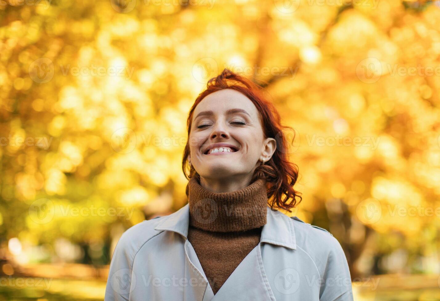 Satisfied caucasian millennial red-haired female in raincoat enjoys warm autumn and freedom, breathes fresh air in park with yellow leaves. Rest and relax alone in city at end of covid-19 quarantine photo