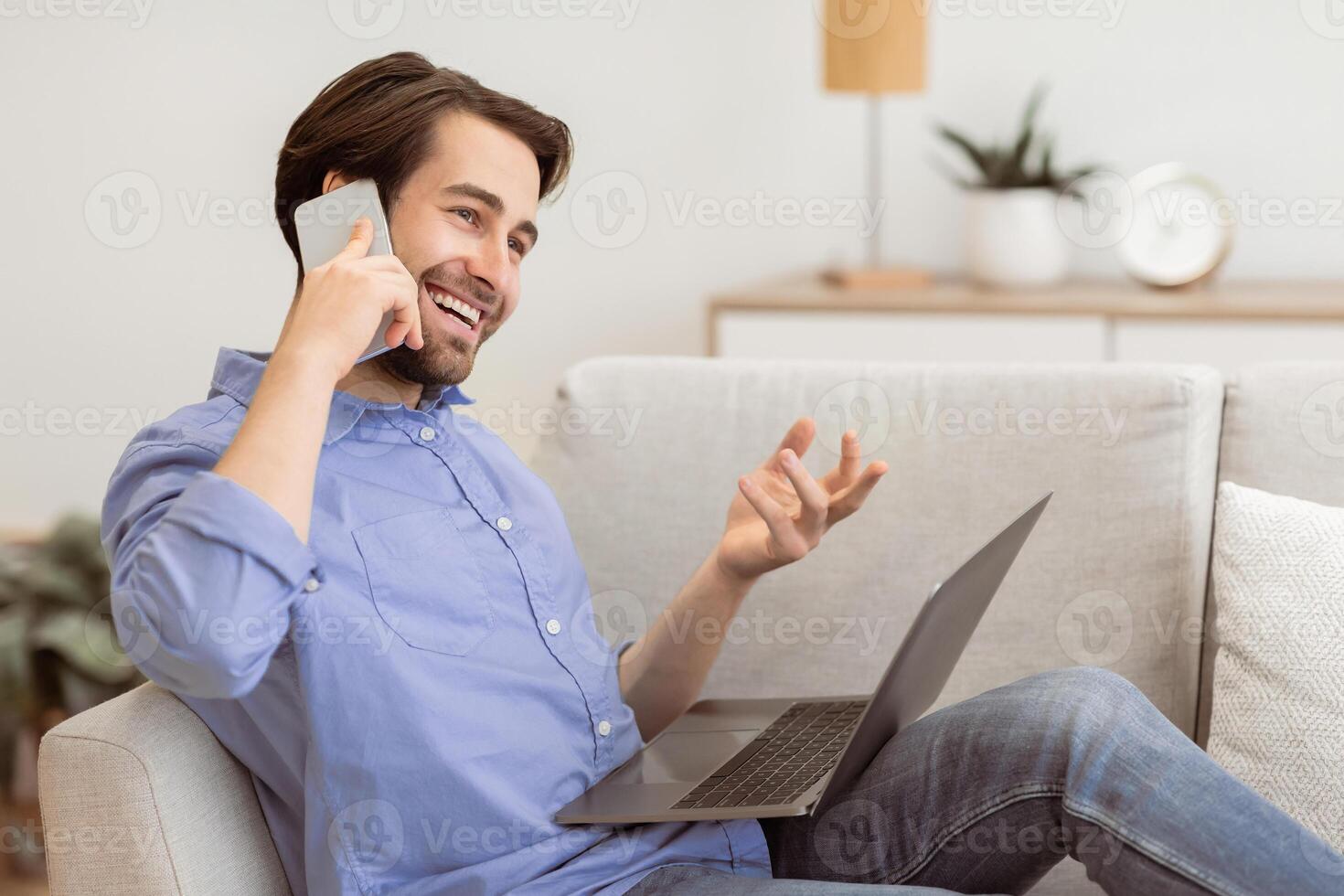 Cheerful Guy Talking On Phone Sitting With Laptop Computer On Sofa At Home. Modern Communication And Lifestyle. Happy Man Chatting On Cellphone And Laughing Indoors photo