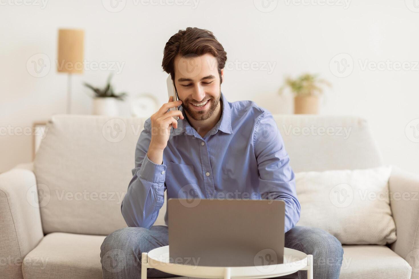 Business Talk. Man Talking On Cellphone Using Laptop Working Remotely From Home, Sitting On Sofa Indoor. Freelancer Speaking By Phone While Works On Computer. Freelance And Distance Work Concept photo
