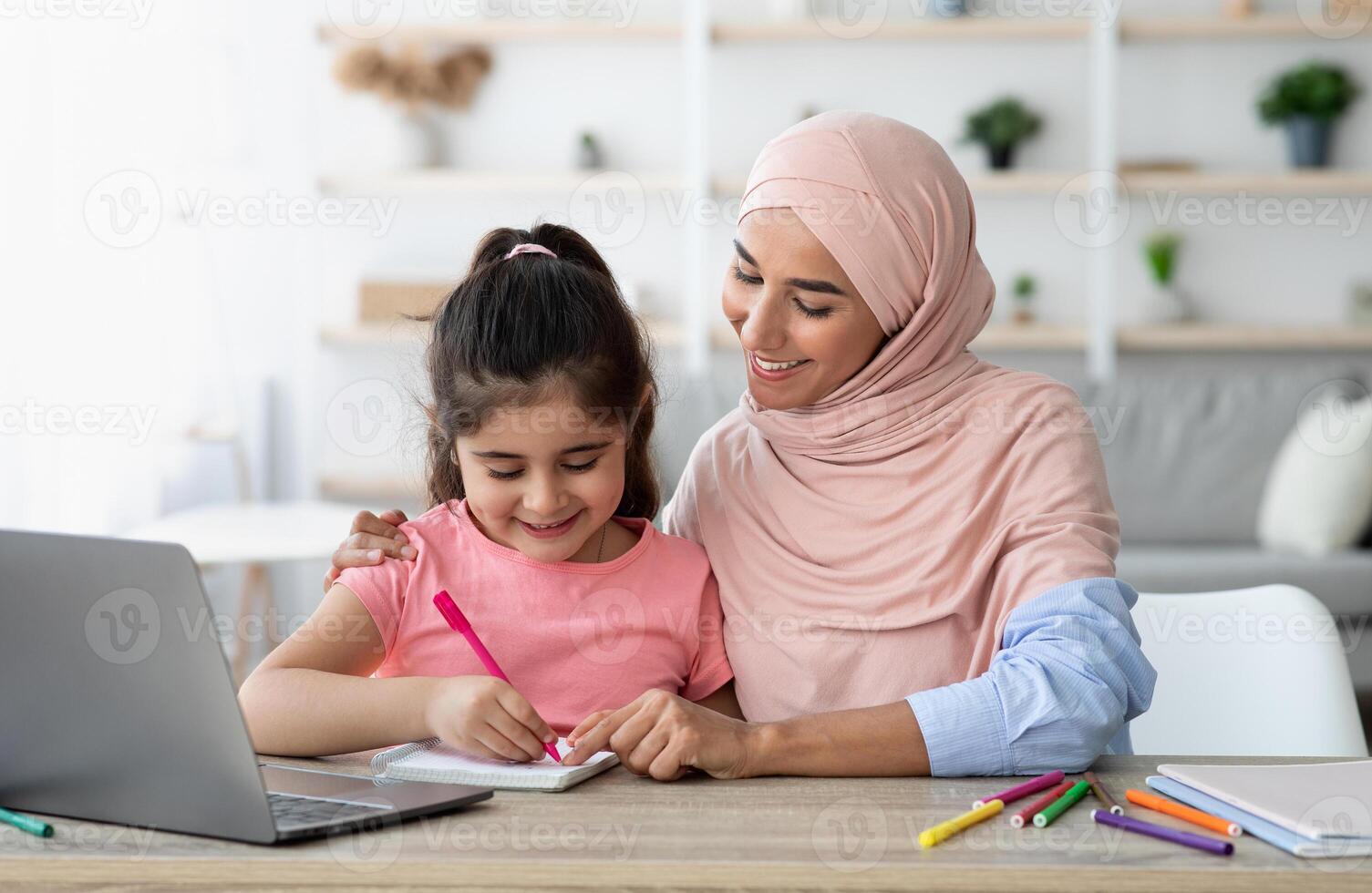 Doing Homework Together. Loving Muslim Mom Helping Her Little Daughter With Study While Sitting Together At Desk With Laptop Computer, Cute Preteen Girl Writing In Notepad And Smiling, Closeup photo