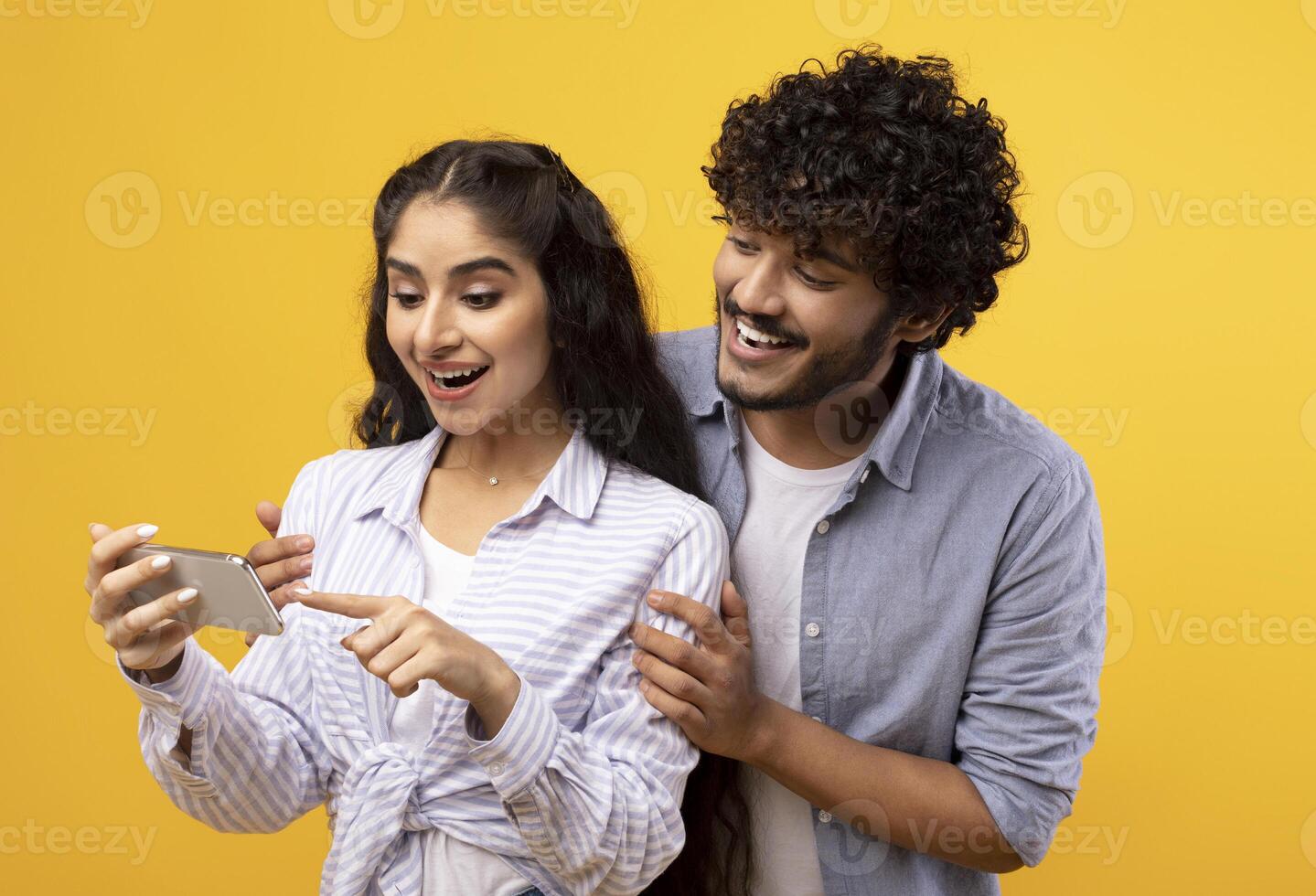 Excited indian couple looking at smartphone screen, using new app or reading cool news, standing over yellow studio background. Happy man and woman using cellphone, guy embracing girlfriend photo