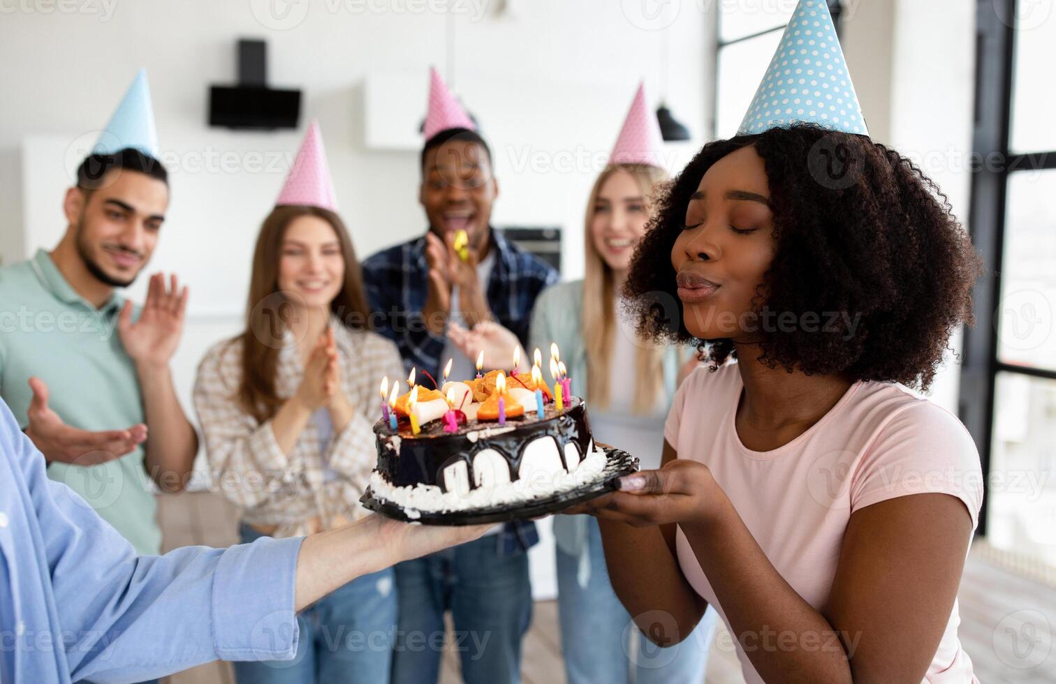 Beautiful black lady blowing candles on birthday cake, closing eyes, making wish, celebrating birthday with her diverse friends at home. Group of young people wearing party hats, having fun photo