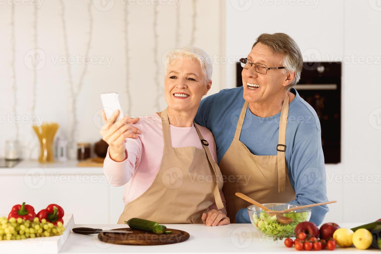 Joyful Elderly Spouses Making Selfie On Mobile Phone Having Fun Cooking Together Standing In Kitchen At Home. Modern Retirement Lifestyle, Senior People Using Gadgets And Enjoying Life Concept photo