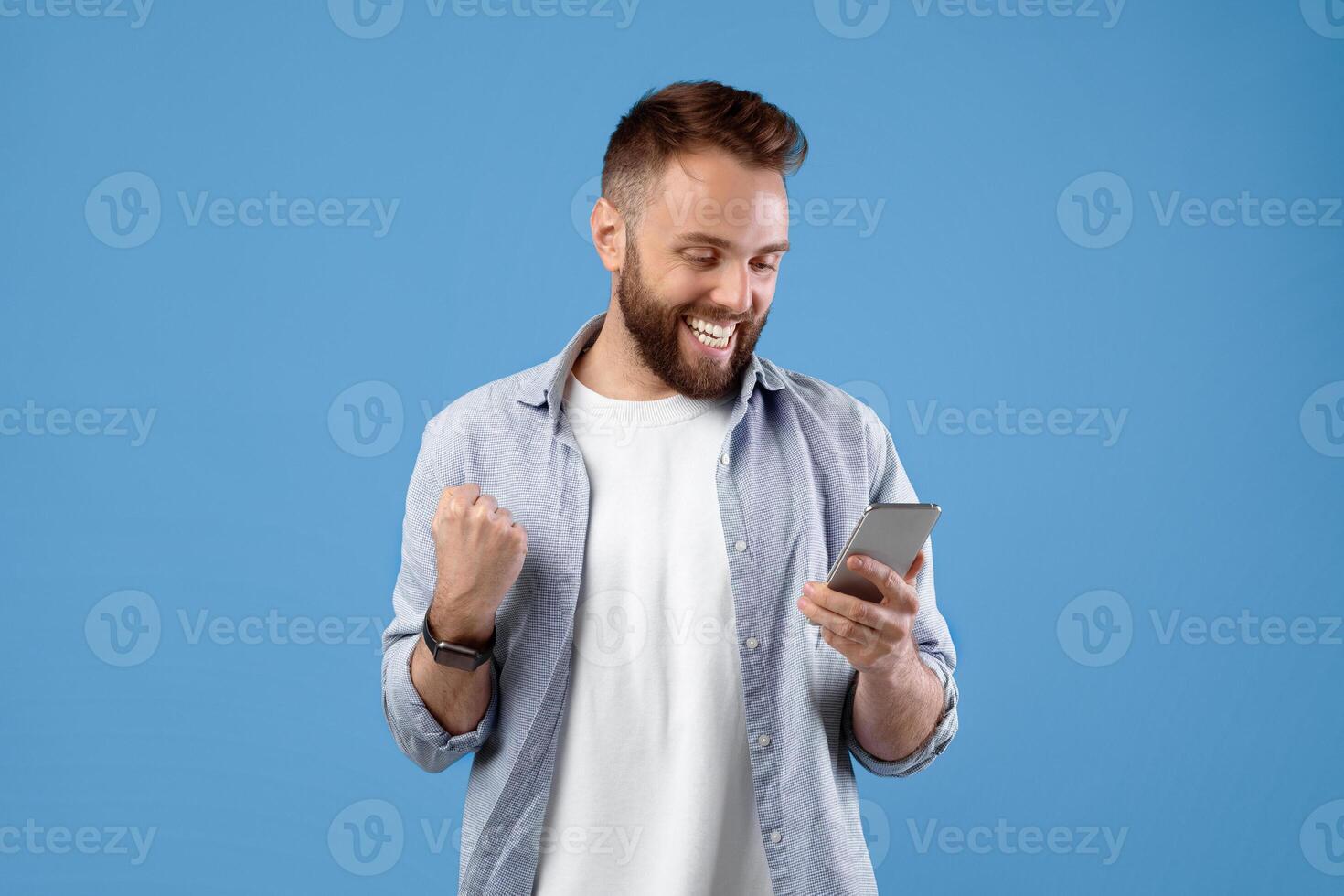 Portrait of happy bearded young man using smartphone, making winner gesture over blue studio background. Excited guy celebrating victory, achieving success, gesturing YES photo