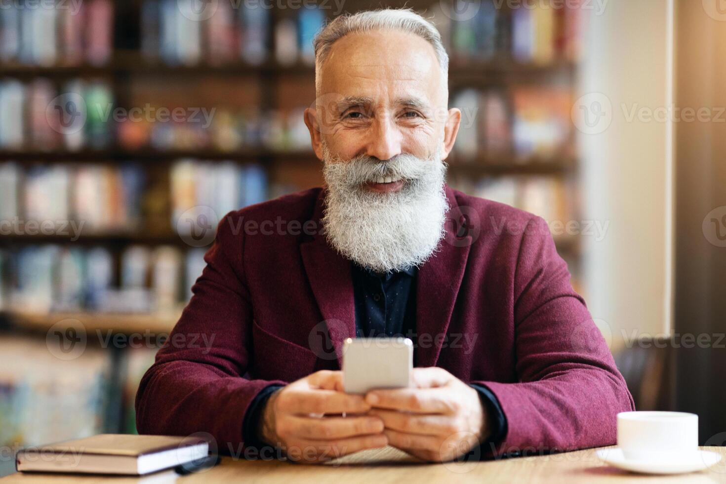 Cheerful elderly grey-haired man with long beard using modern mobile phone while drinking coffee at cafe, chatting with his family or business partners, having notebook by him, free space photo