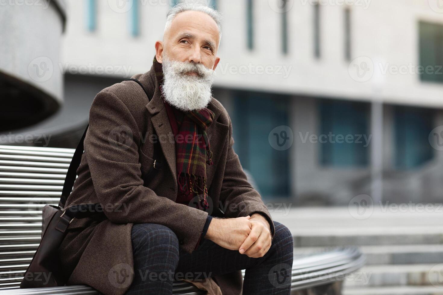 Pensive senior man with grey hair and long beard sitting on bench on the street, wearing warm jacket, having leather bag, resting next to business center after job, looking aside, copy space photo