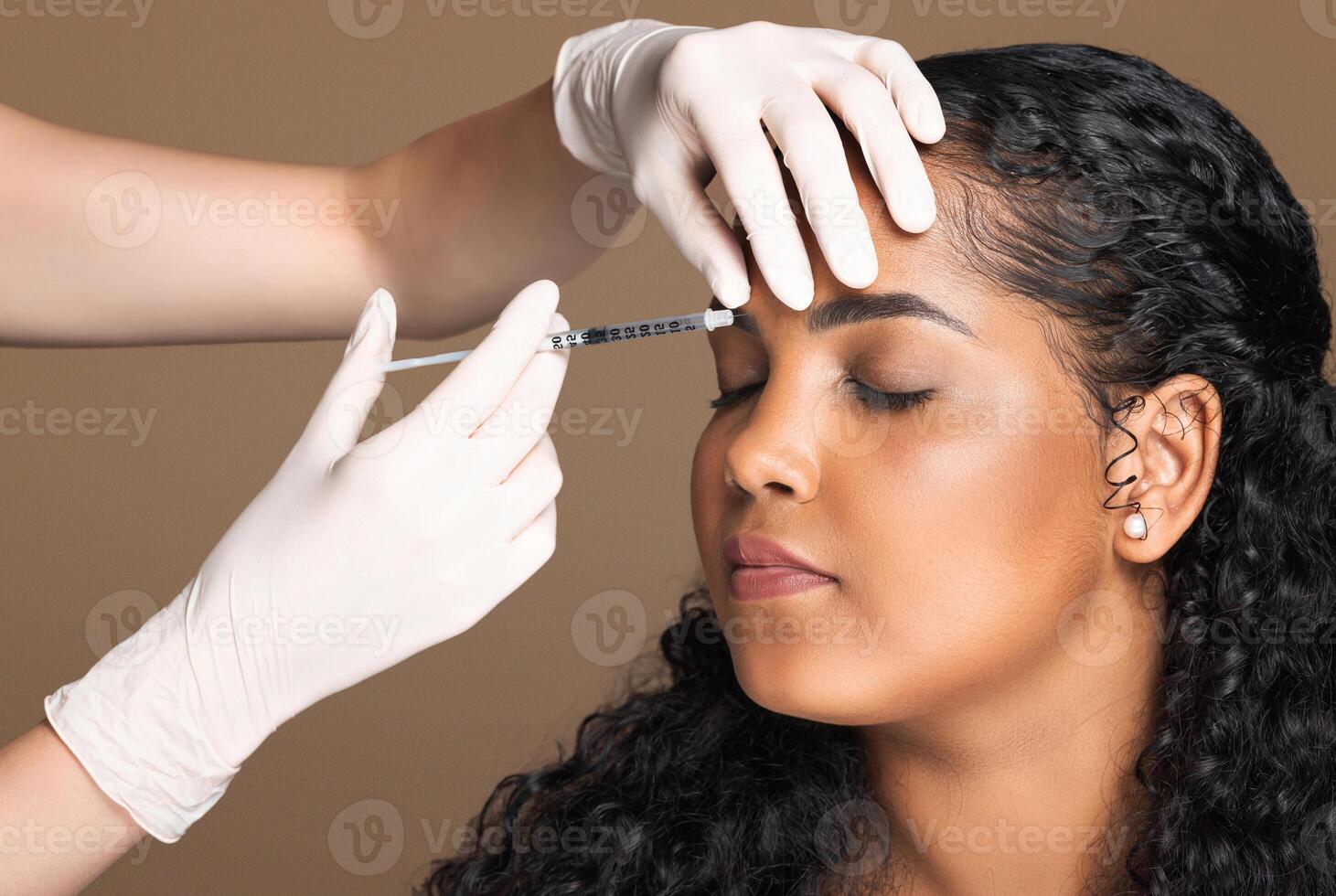 Cosmetologist making botox injection in interbrow zone to latin chubby woman getting anti-wrinke treatment over brown studio background, closeup photo