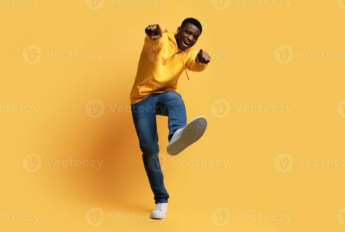 Happy handsome curly young black man in stylish casual outfit showing thumb ups and shoe sneaker sole on yellow studio background, like something or someone, copy space. Human gestures concept photo