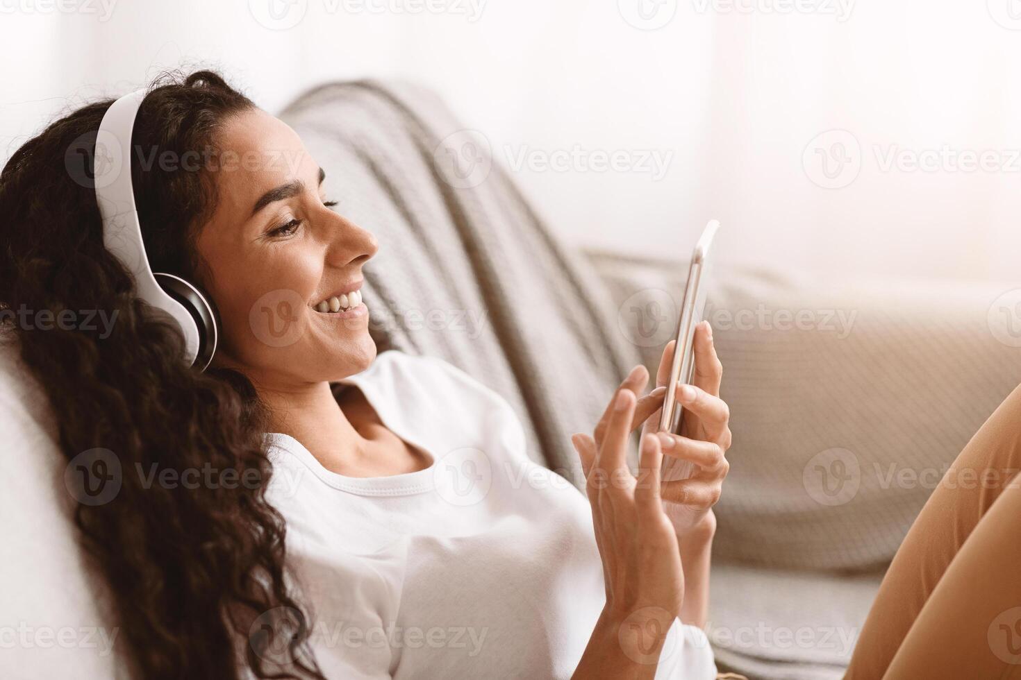 Happy young brunette woman with long curly hair in headphones watching movie or funny videos on mobile phone, using mobile application, sitting on couch at home, copy space, side view photo
