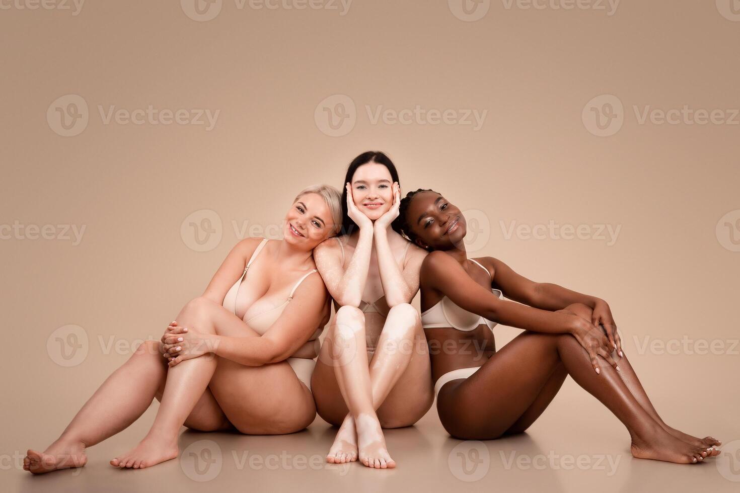 Group of multiethnic women with different body and skin types posing together, attractive multiracial females in underwear sitting on floor over beige background in studio, copy space photo