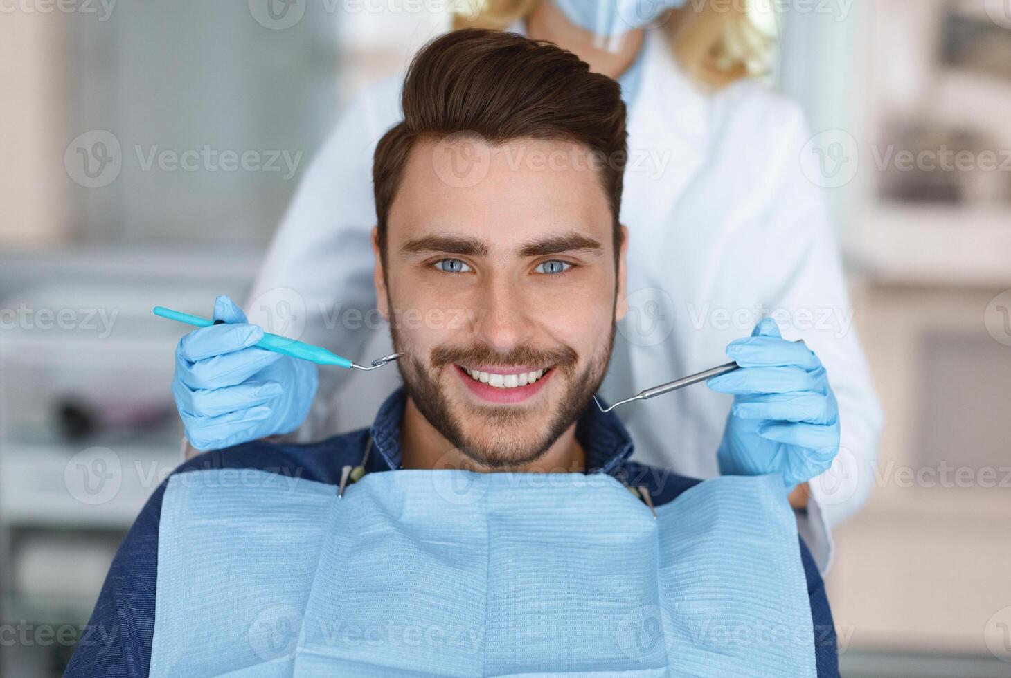 Smiling young bearded man sitting in dental chair, visiting modern dental clinic, having regular checkup, unrecognizable woman dentist holding dental tools next to male patient mouth photo