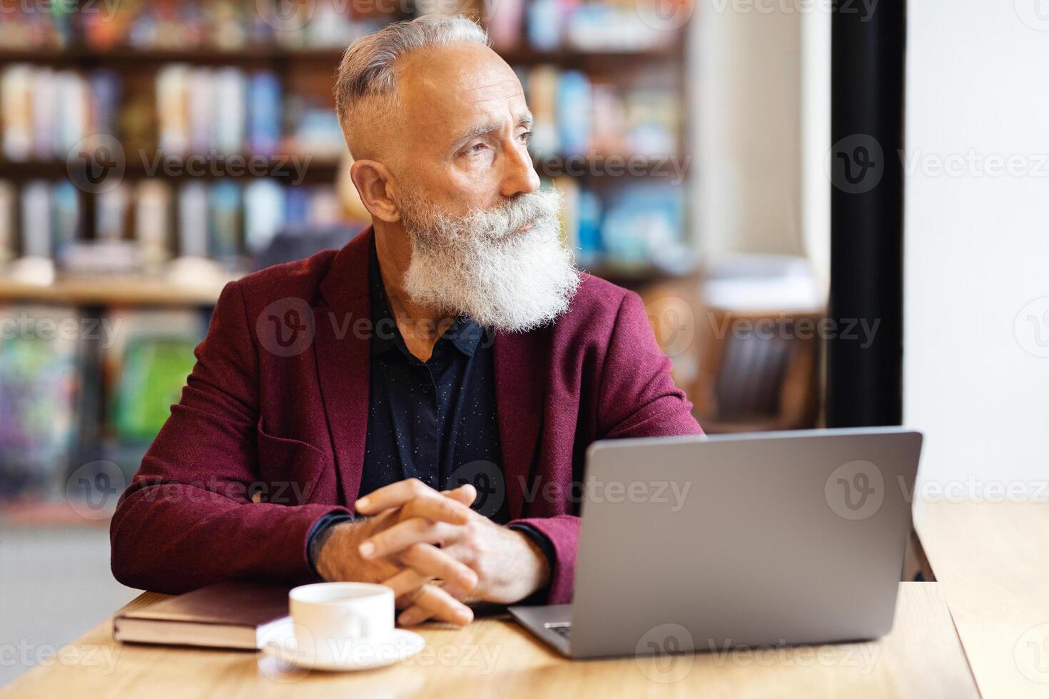 Pensive grey-haired businessman using laptop while having lunch break at cafe, looking through window, copy space. Elderly bearded man working online or websurfing, drinking coffee photo