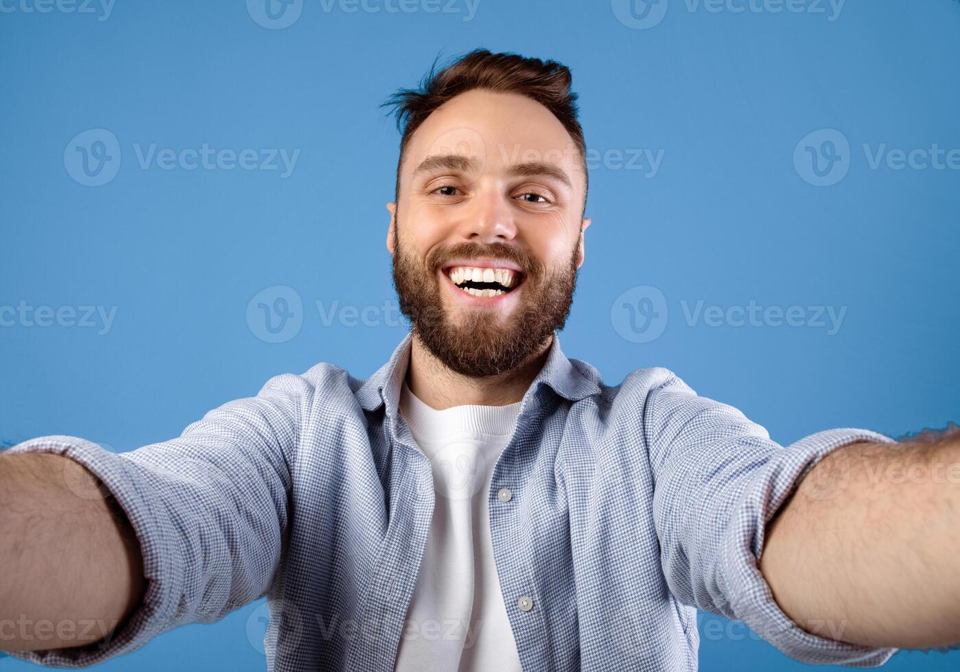 Joyful bearded guy taking selfie on blue studio background. Positive millennial man making photo of himself, smiling at camera, having fun while doing mobile photography