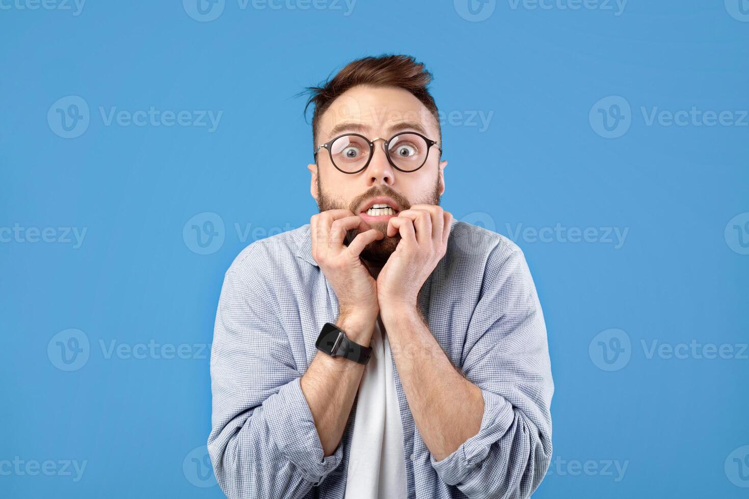 Scared millennial guy biting nails in panic over blue studio background. Horrified young man feeling afraid or nervous, suffering from panic attack, acting dramatic and worried photo