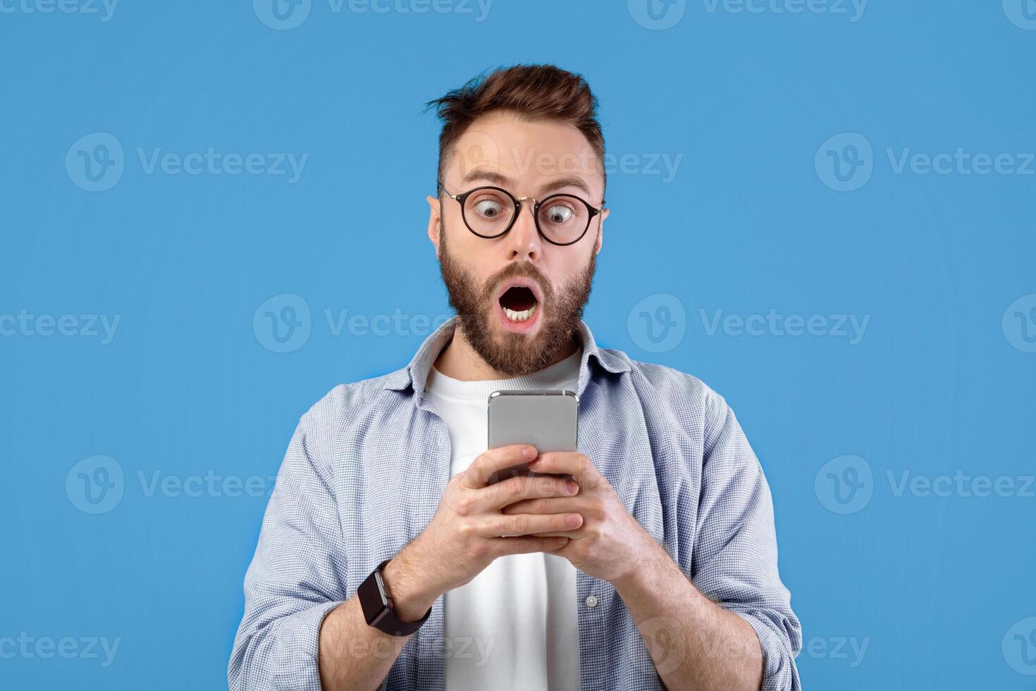 Shocked Caucasian guy staring at cellphone with open mouth over blue studio background, panorama. Surprised young man with mobile device learning unbelievable news, amazed at promo sale photo