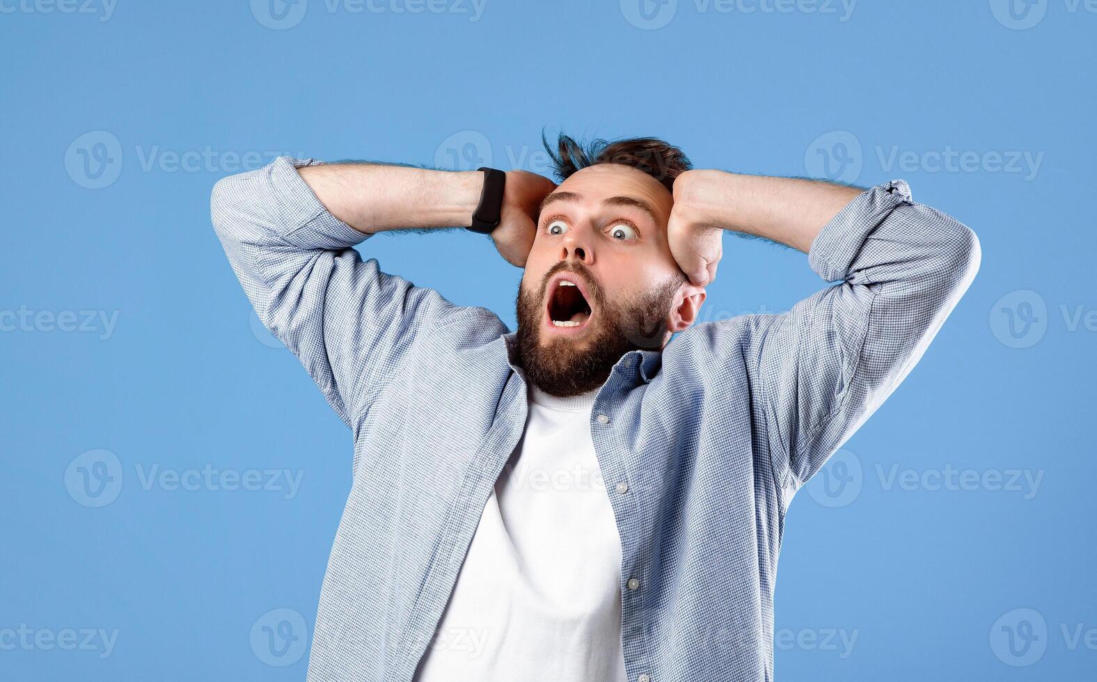 Shocking offer. Extremely surprised young guy shouting OMG in disbelief on blue studio background. Stunned bearded man screaming WOW, cannot believe amazing news, sale, discount photo