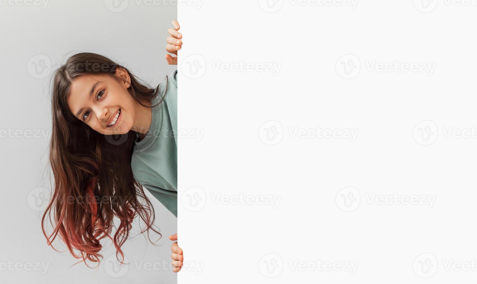 Happy Preteen Girl Posing Hiding Behind Empty White Board And Smiling To Camera In Studio Over Gray Background. Child Holding Paper Poster Advertising Your Text. Panorama, Mockup photo