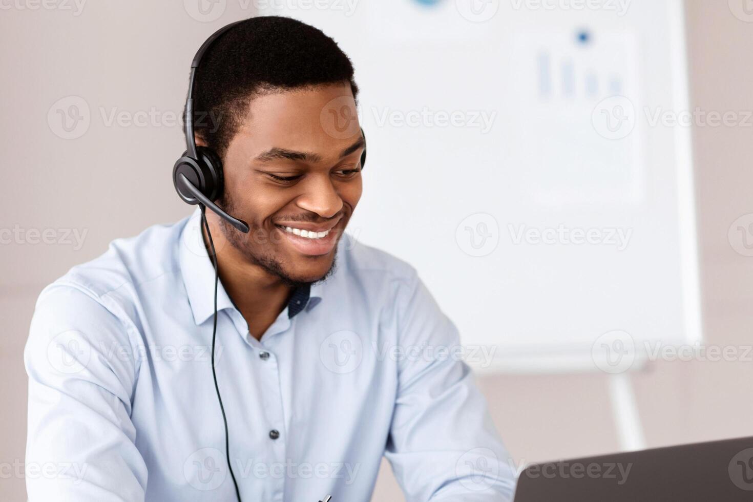 Cheerful black guy crisis manager or customer service representative working in front of laptop in office, wearing headset, having online conversation with clients or business partners, copy space photo