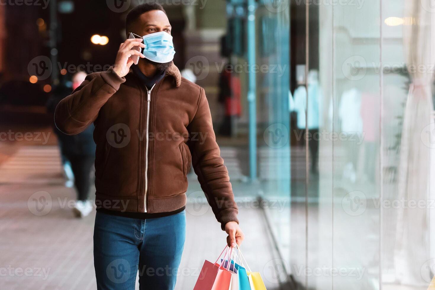 New Normal. Full length of cheerful african american man in medical mask talking on mobile phone, walking down the street in the evening with shopping bags and looking at shop window of the mall photo