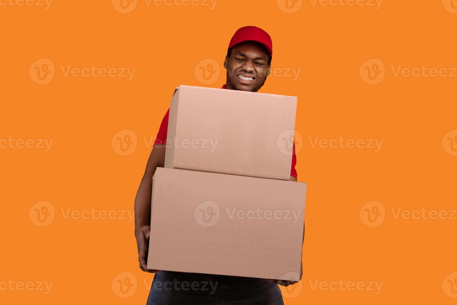 Portrait of african american delivery man wearing red cap and uniform holding stack pile of heavy cardboard boxes, carrying packages with weight stuff, standing isolated over orange studio background photo