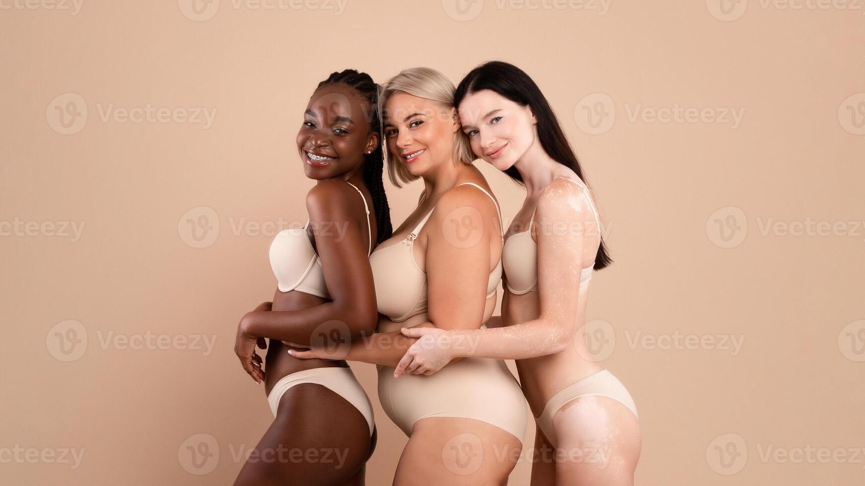 Three multicultural women with different skin and size wearing underwear embracing together, group of beautiful body positive females posing together over beige studio background, copy space photo