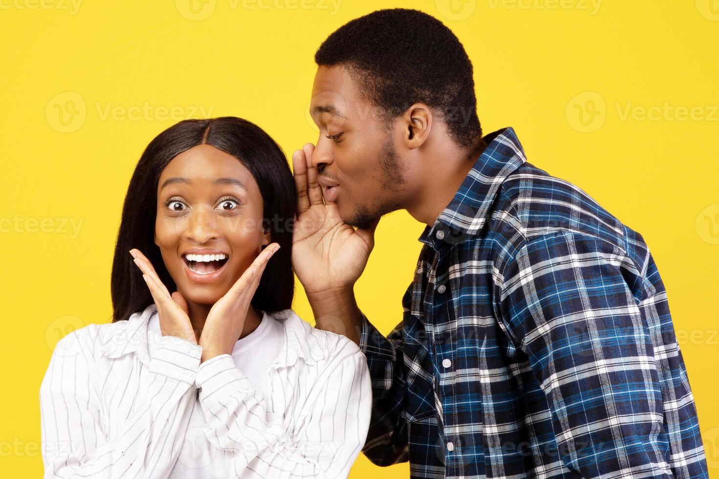 Gossip, secrets and emotions of couple friends. Young african american man speaks in ear of surprised woman in casual, something interesting, isolated on yellow background, studio shot, copy space photo